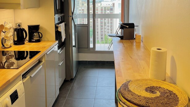 A compact kitchen with modern appliances, a large window with a city view, and a wooden dining table with a woven placemat. - Spacious 2-room apartment with parking in Paris La Défense overlooking the Eiffel Tower   (Puteaux)