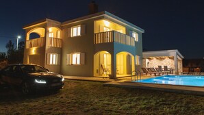 A two-story house with a swimming pool, a car parked in front, and outdoor furniture.