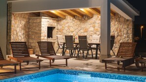 An outdoor pool area with stone walls, wooden lounge chairs, and a table with chairs.