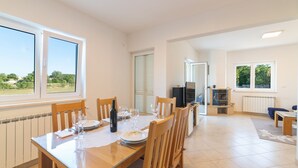 A dining area with a table set for six, a kitchen in the background, and a view of greenery outside.