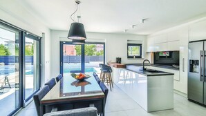 A modern kitchen with a central island, a dining area with a table and chairs, and a large window providing a view of the outdoors. - Modern Kastel Villa with Private Pool, Gym, and Large Outdoor Space - Eight Bedroom Villa (Kastel Novi)