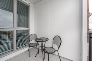 A balcony with a small table and two chairs, a large window with blinds, and a white wall.