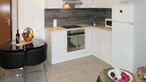 A modern kitchen with white cabinets, a stainless steel oven, a microwave, and a small refrigerator. There is a round table with two black chairs and a basket of fruit. - Sea-View Apartments with Pool, Terrace, and Outdoor Lounge in Scenic Čiovo Island - Two Bedroom Apartment (Ciovo)