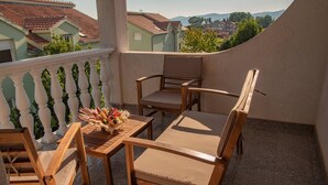 Un balcón con mobiliario de madera, una mesa pequeña con un florero, y vistas a casas y montañas.