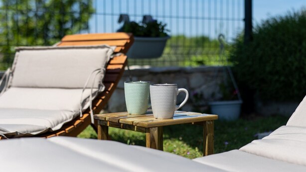 Ein Holztisch mit einer weißen Tasse und einer grünen Kanne, ein Holzliegestuhl und ein hängender Pflanzkasten mit Blumen.