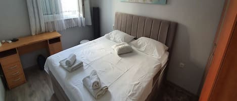 A neatly made bed with white linens and towels, a wooden desk with drawers, a painting on the wall, and a window with curtains.