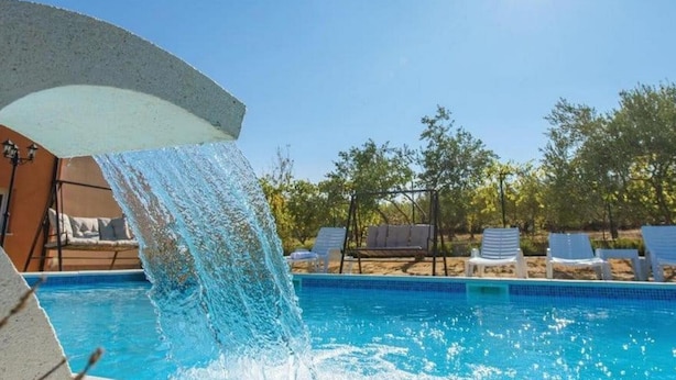 A pool with a waterfall feature, surrounded by lounge chairs and trees.