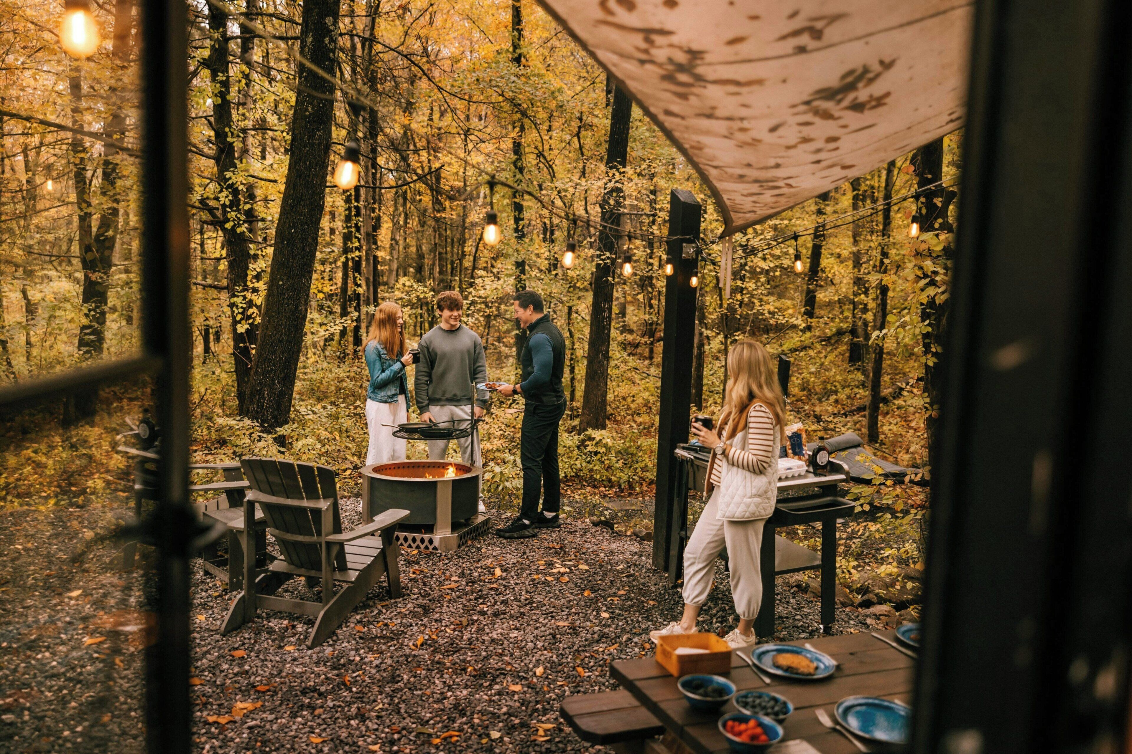 An outdoor dining area with a fire pit, tables, and chairs in a forest setting.