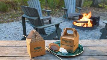 Une table en bois dressée pour un pique-nique, avec des chaises Adirondack, un feu de camp et deux sacs en papier étiquetés « Cabanes à cartes postales » et « S’mores pour deux ».