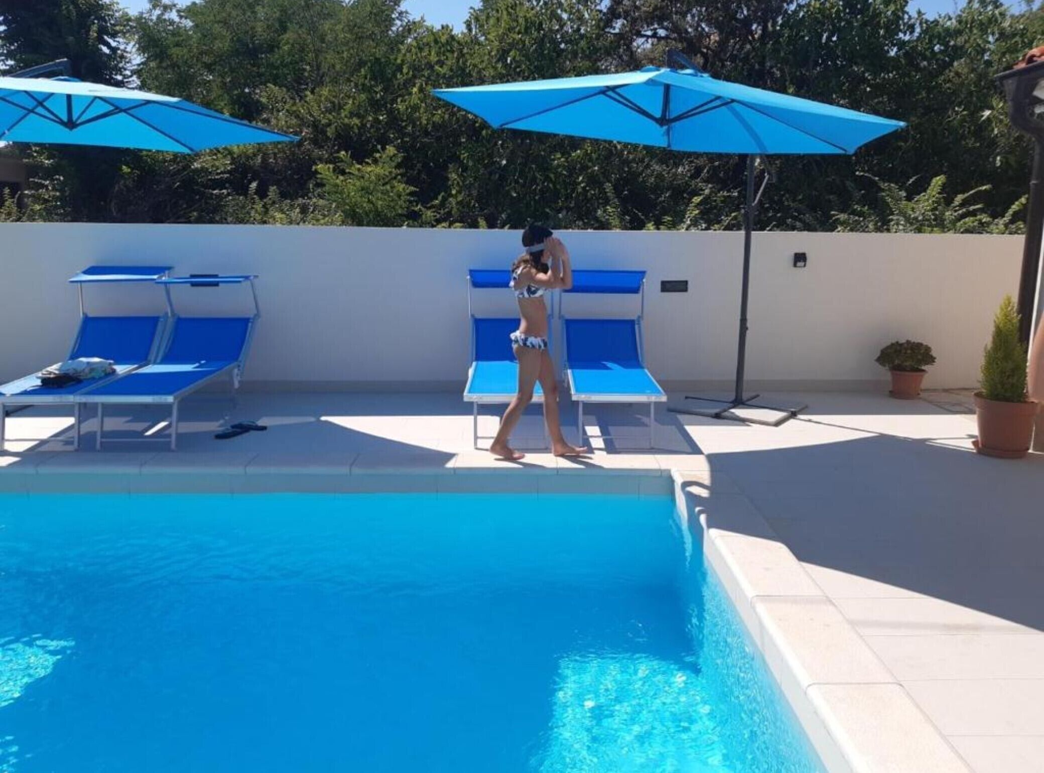 Ein Swimmingpool mit blauen Liegestühlen und Sonnenschirmen, umgeben von einer weißen Mauer und Grünpflanzen.