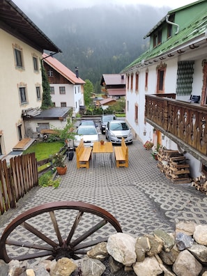 Une cour avec un banc en bois, une table et une grande roue en bois, entourée de bâtiments à l’architecture traditionnelle et de voitures garées.