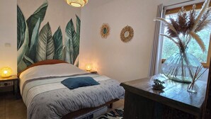 A bedroom with a bed, a nightstand, a wall-mounted lamp, a window with a view, and a decorative vase.