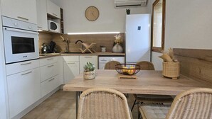 A modern kitchen with white cabinets, a built-in oven, microwave, and a refrigerator. There is a dining table with wicker chairs and a bowl of fruit.