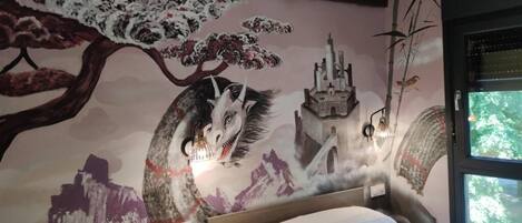 Une chambre avec une fresque représentant un loup et un château, un lit et une fenêtre donnant sur des arbres.