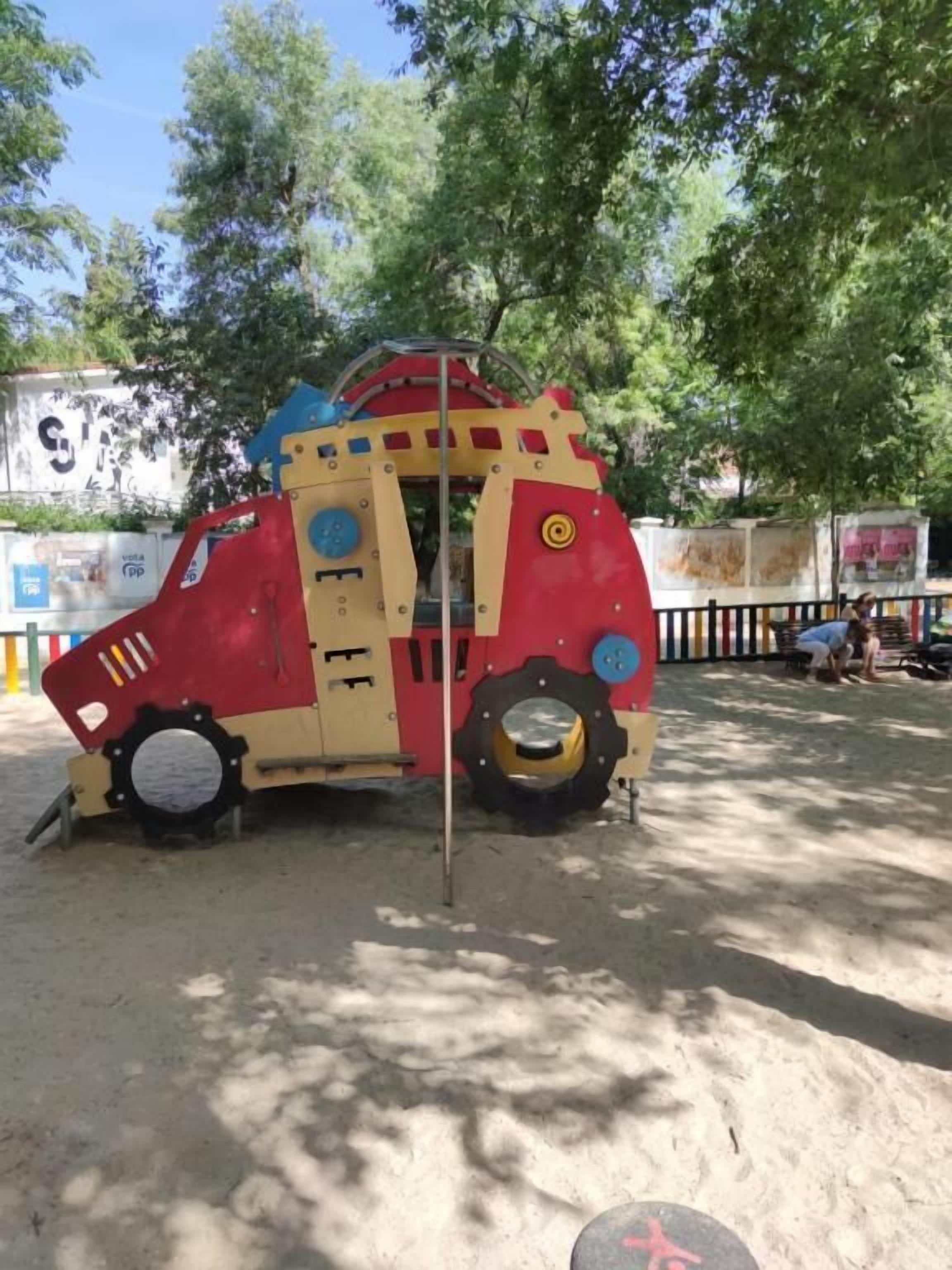 Un vehículo colorido e inmóvil, con diseño rojo y amarillo, posiblemente una estructura de juego o decoración, ubicado en un espacio al aire libre con árboles y una valla de fondo.