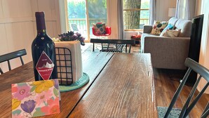 A wooden dining table with a wine bottle and a floral card, a black chair, a grey sofa, and a TV in a cozy living room with a view of the outdoors.