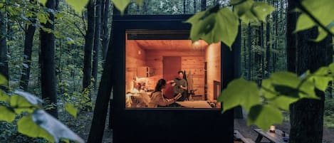 A cabin in a forest with a visible interior including a bed, a table, and a bench.