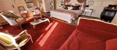 Una habitación con una cama, un sofá rojo, un sillón blanco, una mesita y un espejo.