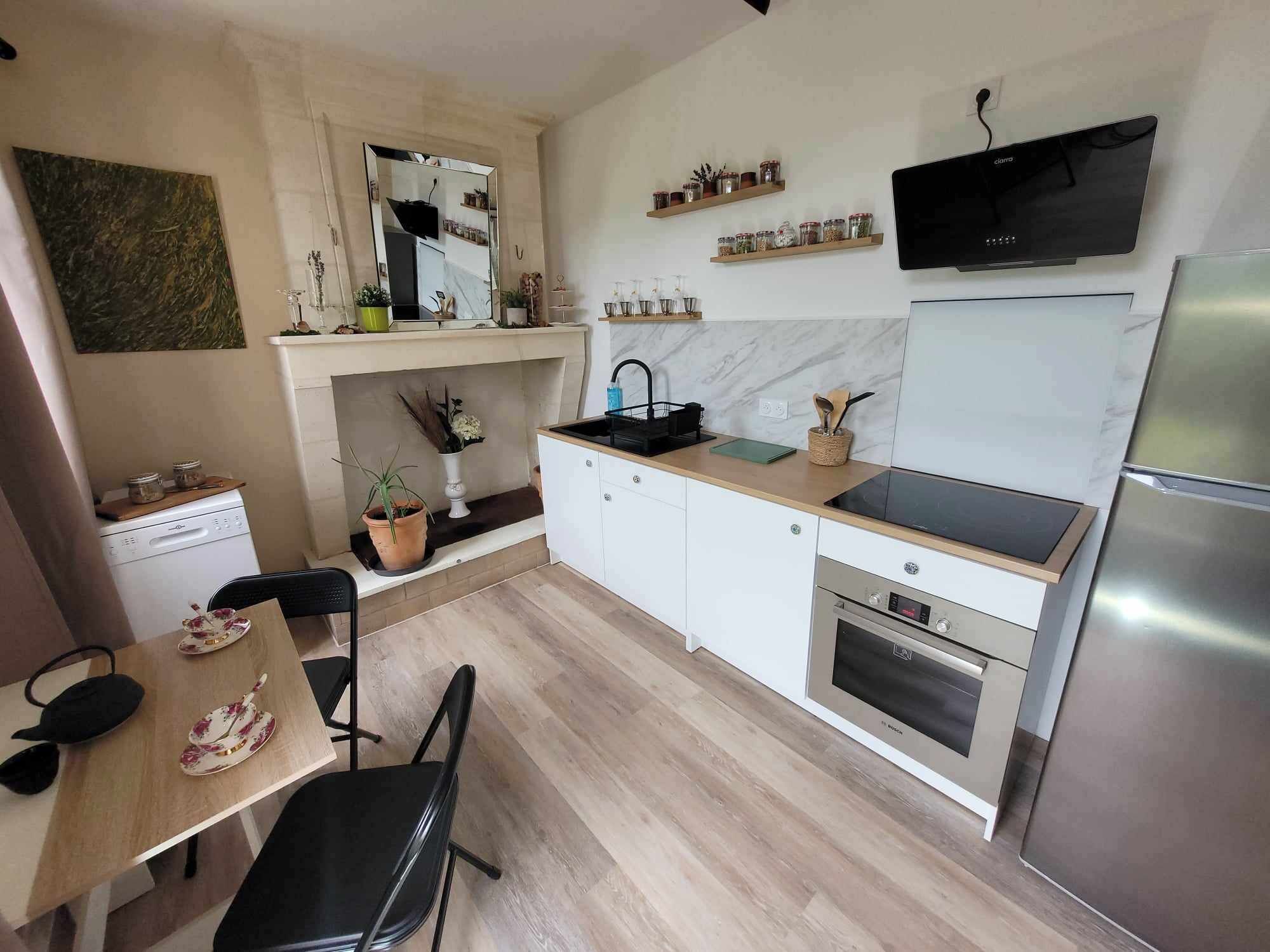A modern kitchen with white cabinets, a black stove, a built-in oven, a microwave, and a stainless steel refrigerator. There is a dining table with chairs, a potted plant, and a mirror above the sink.