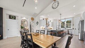 A spacious kitchen with white cabinetry, a wooden dining table, and a large clock on the wall. - Lakefront Retreat: Swim Spa & Backyard Lake Access! Relax & Play; 2 King Suites (Lago Vista)