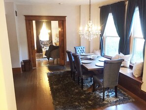 Un coin repas avec une table en bois, six chaises, un lustre et une vue dégagée sur l’extérieur grâce à de grandes fenêtres ornées de rideaux foncés.