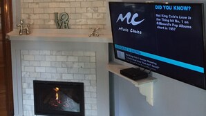 A modern living room with a white brick fireplace, a mounted flat-screen TV displaying a music trivia, and a decorative shelf above the fireplace. - Updated Charming Victorian in Old Town near CSU!  (Fort Collins)