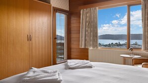 Ein Hotelzimmer mit einem Bett, einem Fenster mit Blick auf einen See und Berge, einem hölzernen Kleiderschrank und einem kleinen Tisch mit einer Lampe.