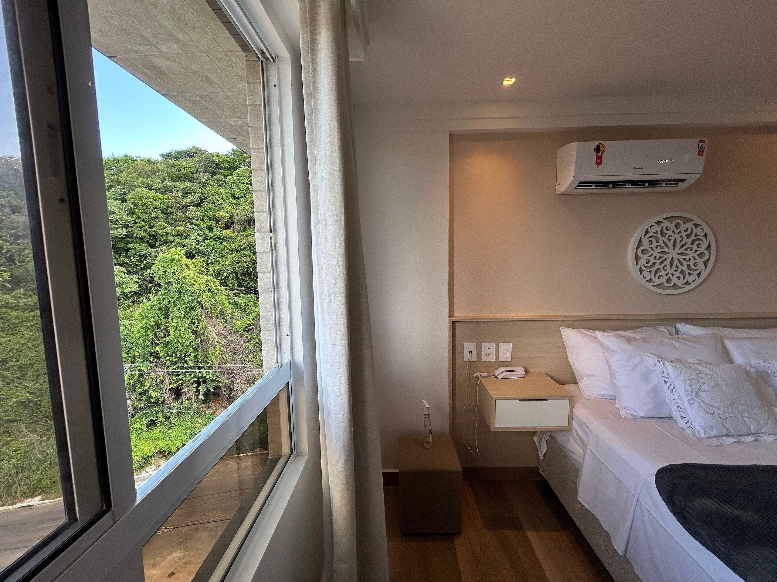 A hotel room with a large window, a bed, a bedside table, an air conditioning unit, and a decorative wall piece.