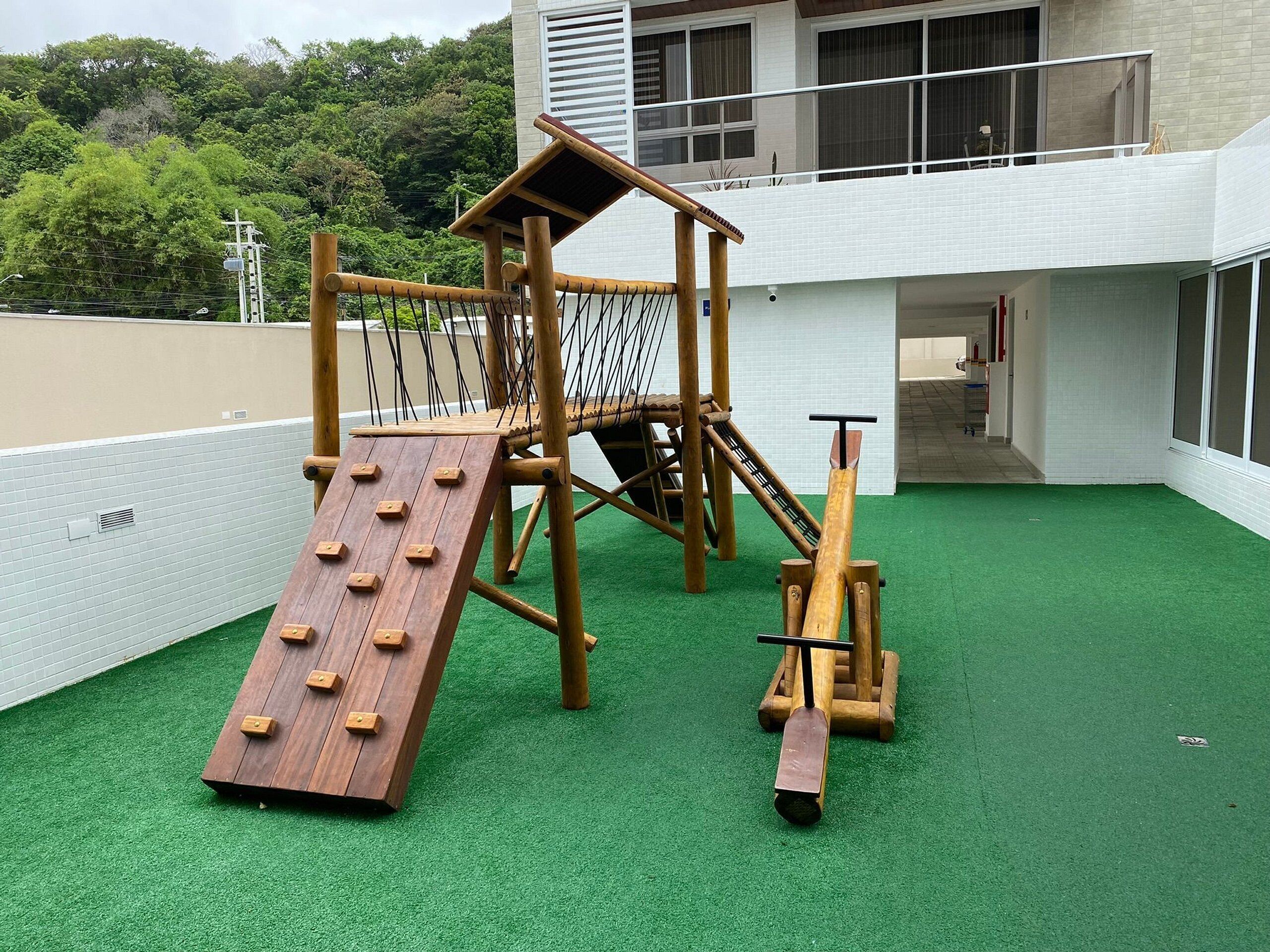 Espaço infantil com escorrega de madeira e estrutura de escalada, cercado por uma grade branca e grama sintética verde.