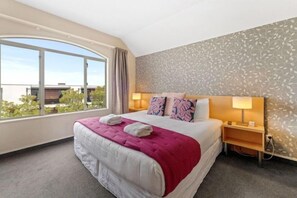Quarto de hotel com uma cama grande, duas luminárias de cabeceira, uma janela com cortinas e um papel de parede estampado.
