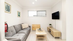 A modern living room with a grey sofa, a wooden coffee table, a TV mounted on the wall, and a window with a lattice design. - Covered Parking | 1 Bdr Apt w/ AC | 435AF1 (Rocklea)