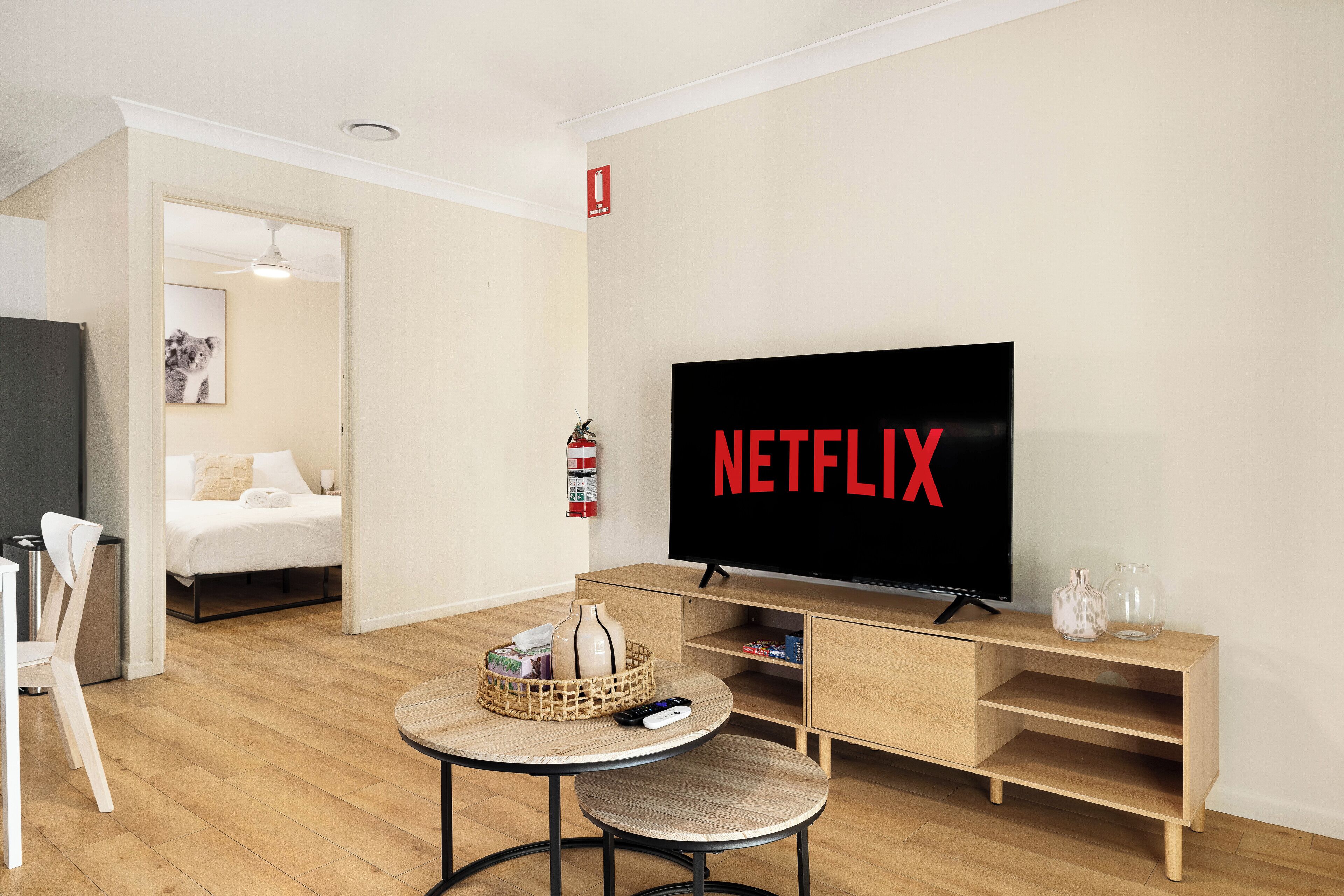 A modern living room with a large flat-screen TV displaying "NETFLIX," a wooden TV stand, a small round table with a basket and remote, and a bed in the background.