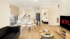Una sala de estar moderna con un sofá negro, un comedor con sillas blancas y una mesa, una cocina con un refrigerador, y un televisor que muestra 