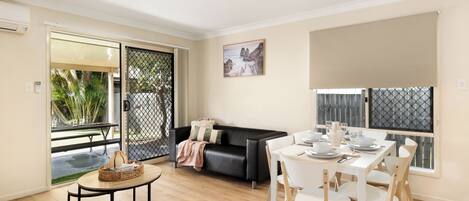 A modern living room with a dining area, a black leather sofa, a round wooden table, and a white dining table with chairs.