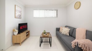 A living room with a grey sofa, a wooden coffee table, a TV on a wooden stand, and a window with blinds. - 1 Bdr Apt w/ AC & Kitchen | Parking | 316F1 (Rocklea)