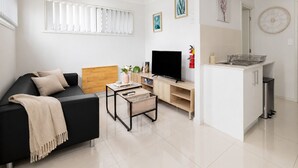 A modern living room with a black sofa, wooden headboard, TV, and a small table with a vase. - 1 Bdr Apt w/ AC | Kitchen | Parking | 131AF1 (Rocklea)