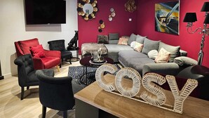 En moderne stue med en rød farve på væggen, en grå sofa, en rød lænestol og en sort lænestol. Der er et fladskærms-tv monteret på væggen, et dekorativt spejl og et billede.