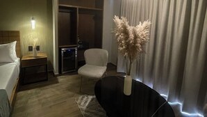 Una habitación de hotel moderna con cama, mesita de noche, silla y mesa de comedor con un jarrón de plumas de pampa.