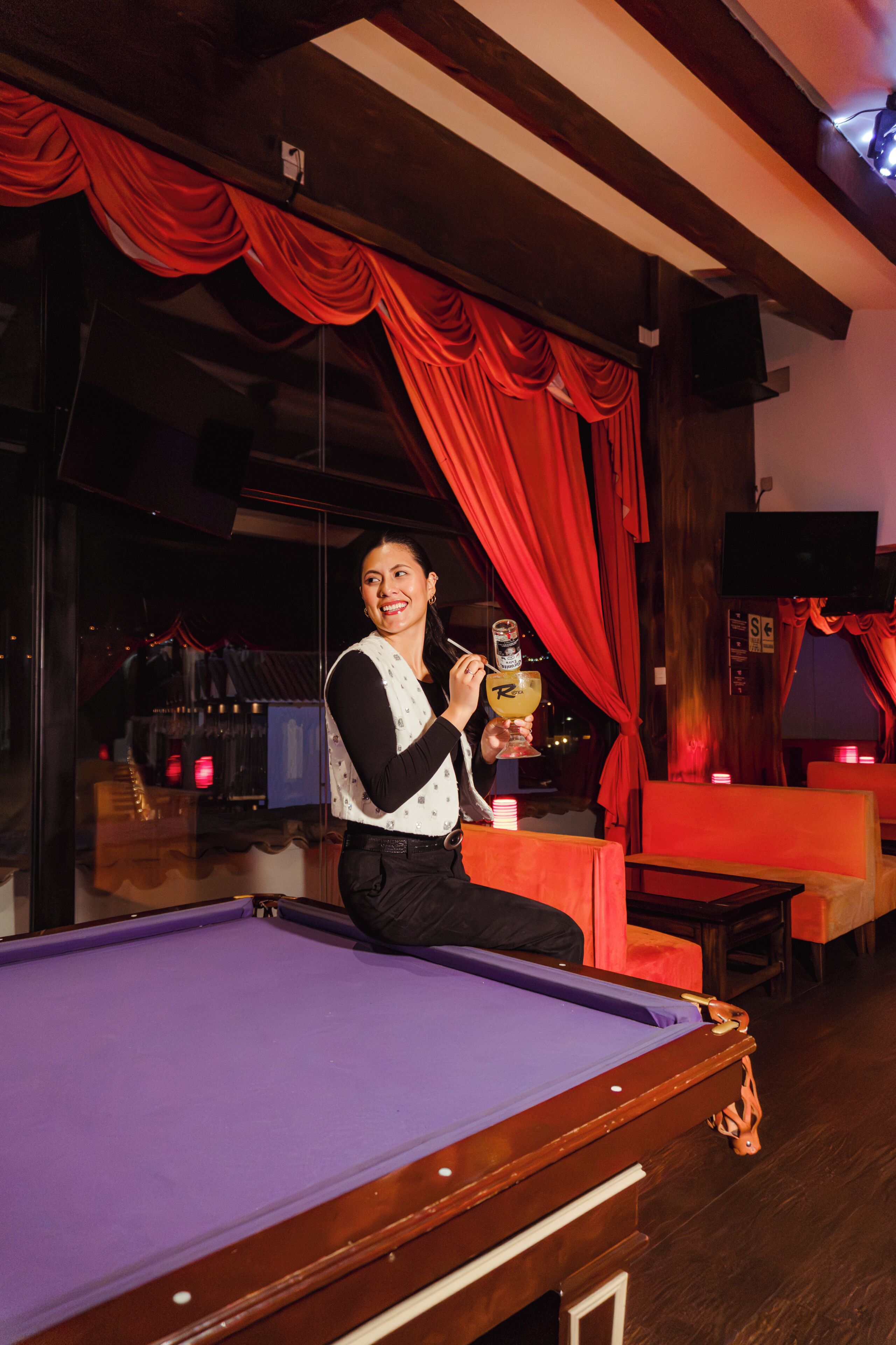 A billiards table, a person holding a drink, and red curtains in a room with wooden beams and a television.