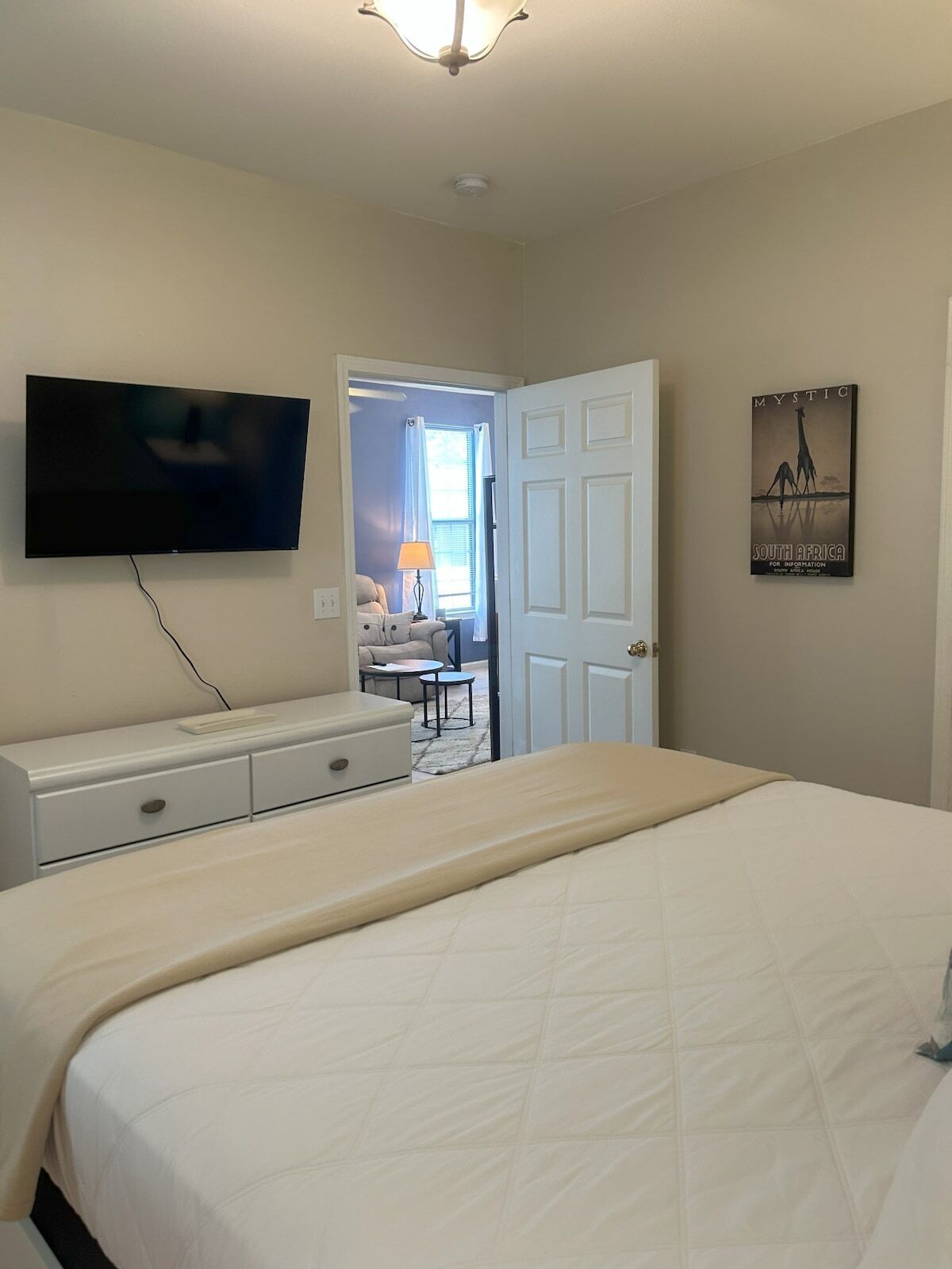 A bedroom with a bed, a TV mounted on the wall, a dresser, a poster on the wall, and a door leading to another room.