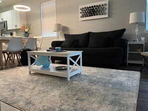 Uma sala de estar moderna com uma mesa de centro branca, sofá preto e uma cozinha ao fundo.
