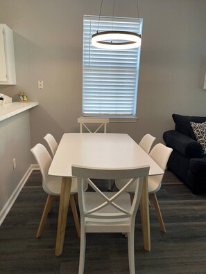 Uma área de refeições com mesa e cadeiras brancas, luminária pendente, janela com persianas e um sofá escuro com almofada estampada.