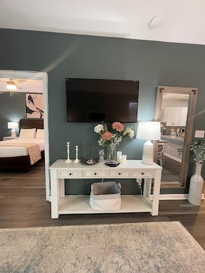 Sala de estar com televisão, mesa console branca com objetos decorativos, cama e espelho refletindo uma área de cozinha.