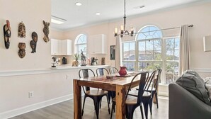 Un espace repas avec une table et des chaises en bois, un lustre et une vue sur l’extérieur grâce à de grandes fenêtres.