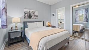 A modern bedroom with a bed, bedside tables, a nightstand, a wall-mounted lamp, and a large mirror. - Captains Quarters In Myrtlewood Villas (Myrtle Beach)