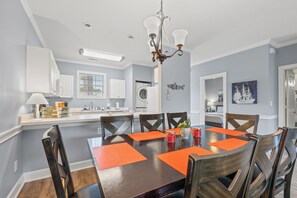 Uma sala de jantar com uma mesa posta para seis pessoas, rodeada por cadeiras de madeira, um lustre e uma cozinha ao fundo.