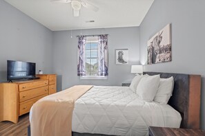 Une chambre à coucher avec un lit, un téléviseur posé sur une commode en bois, une fenêtre avec des rideaux et un ventilateur de plafond.