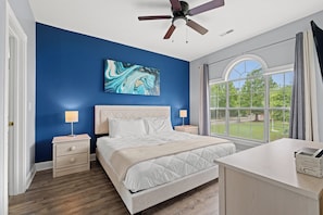 A bedroom with a bed, two bedside tables, a ceiling fan, a large window with curtains, and a wall painting.