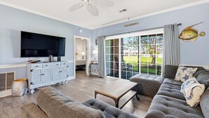 A living room with a flat-screen TV, grey sofas, a wooden coffee table, a white ceiling fan, and a large window with curtains.