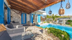 Zona junto a la piscina con mobiliario de mimbre, pared de piedra y pérgola de madera.